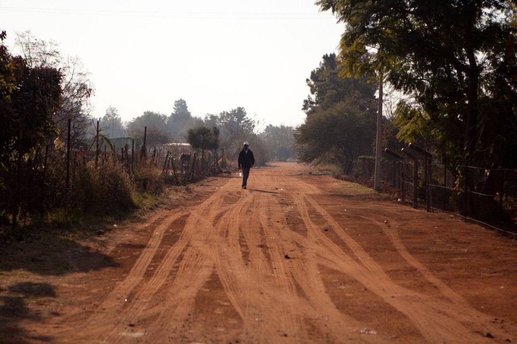 An South African man walks down a dirt road with trees on the right side and an flimsy fence on the left