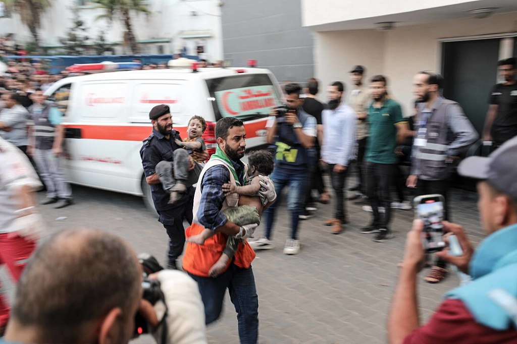 Two men rush past a crowd carrying one child each. In the background is an ambulance, in the foreground another man from the crowd films them on his phone. It is a scene from the early days of the Gaza Genocide.